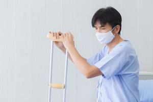 Asian patient male who wearing a medical face mask and holding the crutch walking due to his leg broken is admitted to the hospital in Corona virus Covid-19 pandemic situation. photo