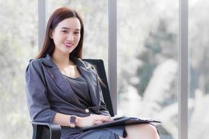 Professional Asian business woman in dark grey dress sits on chair in office room and works happily with a mirror as a background. photo