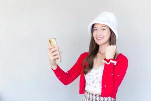 Beautiful young Asian woman who wears a red coat and white hat as a Santy girl holds a smartphone in her hand and another act while looking at the camera as glad and cheerful smiling. photo