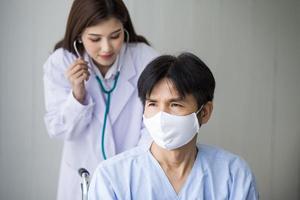Asian woman doctor use a stethoscope to check the lung rhythm of a male patient who wear face mask while he sit on a wheelchair at hospital. photo