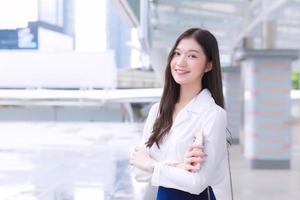 Asian confident working woman who has a long hair with a white shirt is arm crossing in hand holding a smartphone standing urban outdoors while walking to office in big city with business buildings. photo