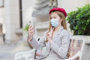 Asian female wears suit and holds smartphone for video call in her hands. She acts a two thumbs up sign showing victory while sits on bench in park outdoors while wearing a medical face mask photo