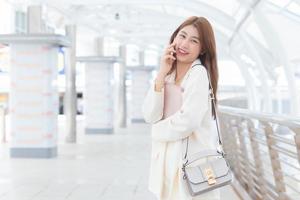 una trabajadora profesional asiática con traje blanco sonríe y llama a los clientes en la oficina mientras sostiene una tableta y una bolsa en sus manos en un feliz día de trabajo con edificios comerciales.