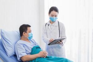 Asian woman doctor talking with a man patient who he is on bed about his pain and symptom in hospital. They wear a medical face mask to protect respiration system Covid-19. photo