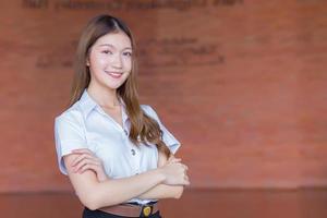 Portrait of adult Thai student in university student uniform. Asian beautiful girl standing smiling with her arms crossed on a brick background. photo