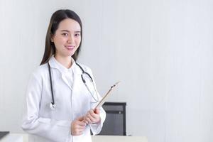 Professional beautiful young woman doctor holding document in clipboard smiling looking at the camera while she wears a white lab coat and a stethoscope in hospital. Health concept. photo