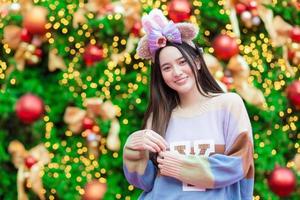 A beautiful Asian woman in a colorful sweater stands smiling and happy in front of the Christmas tree With bokeh as a background in the theme to celebrate Christmas and Happy New Year photo