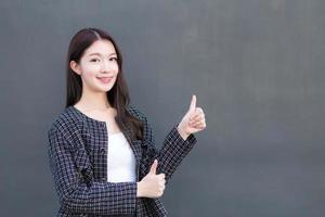 Asian professional working woman who wears black suit with braces on teeth is pointing hand to present as thump up on the dark gray wall. photo