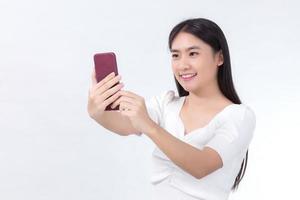 Portrait of Asian beautiful woman who has black long hair in white shirt, is holding the smartphone in her hand and smiling. She take a selfies photo on white background.