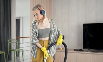 Young girl havig fun while cleaning floor with vacuum cleaner. Happy woman doing housework at home enjoy music wearing earphones. photo