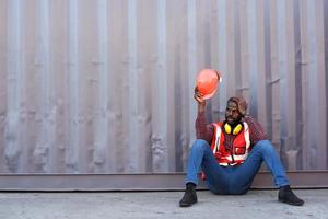 African American male technician or engineer. Sit near a container and look tired and sleepy or unemployed. Logistics industrial cargo mover concept. photo