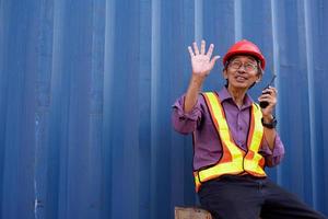 A senior elderly Asian worker engineer wearing safety vest and helmet standing and holding radio walkies talkie at logistic shipping cargo containers yard. photo