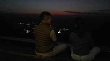 vue arrière de la silhouette d'un couple asiatique assis sur une terrasse parlant et profitant de la vue sur la nature au coucher du soleil video