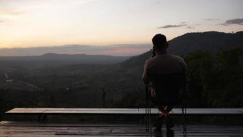 Rückansicht eines Mannes, der mit dem Smartphone auf der Terrasse sitzt und den Sonnenuntergang betrachtet video