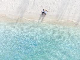 Pareja joven viajera sentada y relajada en una hermosa playa tropical de arena blanca con espuma de olas y mar transparente, vacaciones de verano y vista superior de fondo de viaje desde un dron foto