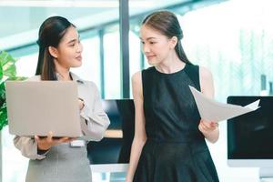 Two young business woman discussing project and report in laptop and digital tablet while standing and analyzing data on statistics and brainstorming information in modern office photo