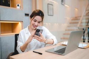 sonriente joven mujer de negocios asiática y empresaria usando teléfono móvil y desplazándose y navegando a través de las redes sociales tomando un descanso después de trabajar en una computadora portátil foto