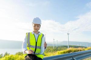 Beautiful caucasian woman in white helmet looking through some documents on clipboard while standing on farm with windmills. photo