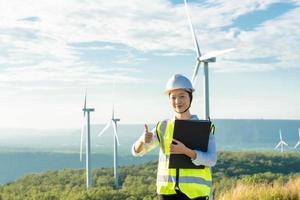 Pretty caucasian woman smiling and looking at camera with document in hands. Female person wearing formal clothes, white helmet and safety glasses for work on windmill farm. photo