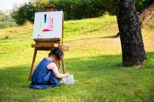 A little girl is sitting on the grass and painted on the canvas placed on a drawing stand photo