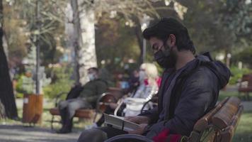 Dark-skinned teenager reading a book in the park. Reading a book in the park. Drinking coffee while reading a book. Reading a book outdoors. pandemic conditions. The man in the black mask. video