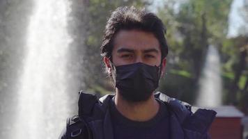 Portrait of teenager in black mask. Brunette student looking at camera. Shoulder background shot. Water fountain in the background. Natural life in the park. video