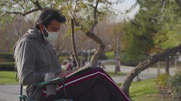 giovane seduto sulla sua sedia nel parco e leggere un libro. leggere un libro nel parco. fare una pausa libro nella natura. condizioni pandemiche. maschera bianca. bere caffè. girare pagina. video