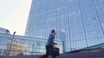 hombre de negocios tarde al trabajo, edificios de lujo. el hombre de negocios que llega tarde al trabajo mira su reloj y corre hacia el edificio. hombre de negocios en traje con bolsa. video