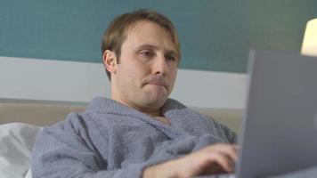 Man working on laptop while lying in bed. Man lies on bed in bathrobe, works on laptop, thoughtful. video