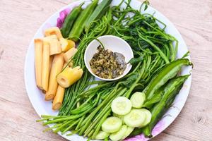 Verduras al vapor en plato y fondo de madera, comida tailandesa asiática con verduras al vapor o hervidas con brotes de bambú de pepino acacia pennata okra frijol alado ceilán espinacas y salsa de chile foto