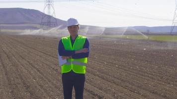 ingegnere guardando la fotocamera in terreni agricoli. l'irrigazione viene effettuata nel terreno agricolo. ingegnere che tiene tablet guardando la fotocamera e ridendo. video