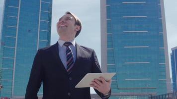 hombre de negocios alegría de éxito. el hombre de negocios se regocija trabajando en una tableta frente a edificios comerciales de lujo. Hombre de negocios exitoso video