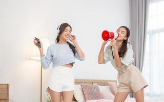 Pair of beautiful Asian female friends happily dancing and singing with a megaphone in the bedroom. photo