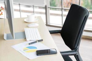 Business and workplace concept. Cup of hot coffee on work desk with computer keyboard Mobile and papers graphs in office working room. photo