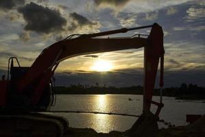 las excavadoras están estacionadas después del trabajo por la noche, usando los brazos del camión como marco para ver el hermoso cielo y las nubes de varios colores por la noche, un rayo de luz en el hermoso río foto