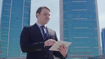 hombre de negocios que trabaja con tableta frente a edificios comerciales. hombre de negocios en traje trabaja con tableta frente a edificios comerciales modernos. video