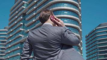 hombre de negocios hablando por teléfono frente a edificios comerciales. hombre de negocios en traje hablando por teléfono frente a modernos edificios de negocios. video