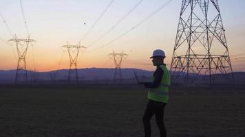 ingeniero caminando entre torres eléctricas. ingeniero con laptop en mano camina entre postes de energía e inspecciona. toma notas en la laptop y mira los polos. video