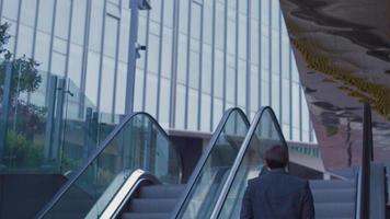 hombre de negocios con bolsa subiendo la escalera mecánica. hombre de negocios con maletín frente a un edificio de negocios de lujo sube la escalera mecánica. imagen de edificio moderno y hombre de negocios moderno. video