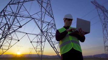 elektrotechnisch ingenieur die voor hoogspanningslijnen werkt. ingenieur die bij zonsondergang aan laptop werkt en naar elektrische palen kijkt. video