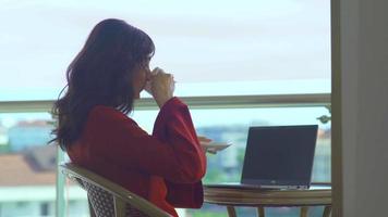 empresaria tomando café en el balcón del hotel. mujer sentada en el balcón del hotel con vista al mar y sosteniendo una computadora portátil frente a ella tomando su café. video
