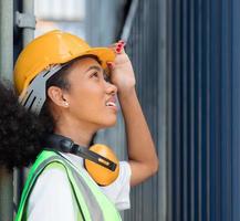 Beautiful African American female foreman worker wears orange safety hardhat watching cargo loading boxes at container yard, portrait. Industrial engineer black woman works at international dockyard photo