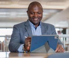 retrato de un hombre de negocios negro feliz con traje sentado en un escritorio en una oficina moderna usando una tableta para una reunión remota. gerente negro sonriente, trabajando con éxito como trabajador de cuello blanco en la empresa. foto