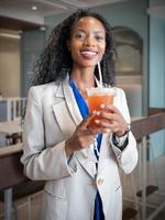 Portrait of black woman with long afro hair style standing and holding cold beverage looking at camera smiling. Businesswoman enjoys relaxing with glass of caffeine at coffee break for refreshment. photo