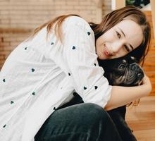 Charming young woman lying on the floor, looking at the camera smiling with her adorable french bulldog stepping on her back. Cheerful girl enjoying, bonding with her funny dog. Pet lover lifestyle. photo