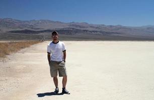 Young man with sunglasses and shorts standing in Death Valley next to a road winding its way through the mountains in the background photo