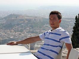 apuesto joven con una cara seria sentado al sol con la ciudad de atenas, grecia, en el fondo foto