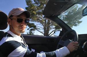 Young man with wooden sunglasses and baseball cap driving a convertible