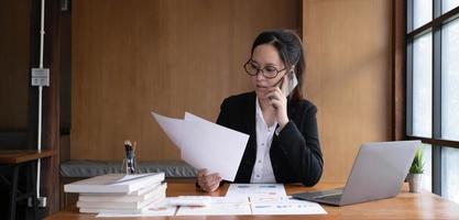 Serious asian businesswoman using laptop looking at computer talking on phone consult client sit at desk, focused female sales manager make mobile business call discuss work with customer in office photo