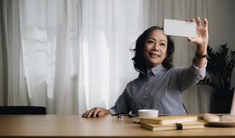 Asian senior elderly woman smile and video call in living room at home. Strong elderly older grandmother feeling happy using mobile cell phone communicate with family enjoy retirement life in house. photo